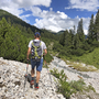 Wanderer mit Rucksack auf einem Bergweg in alpiner Landschaft mit Bäumen und Fluss.