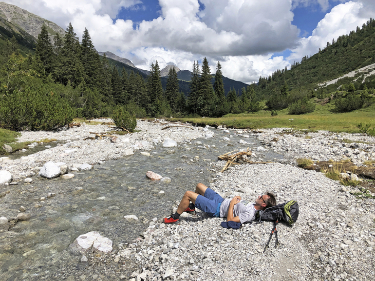Wanderer rastet am Flussufer in alpiner Landschaft, umgeben von Tälern und Wolken.