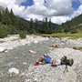 Wanderer rastet am Flussufer in alpiner Landschaft, umgeben von Tälern und Wolken.