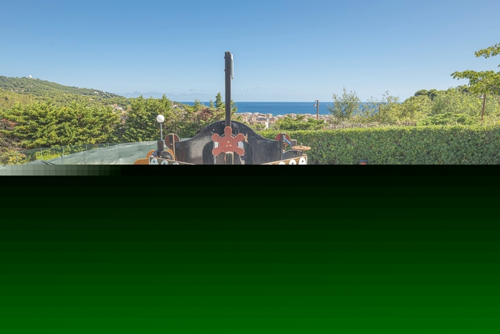 Spielplatz im Freien mit Kletterschiff und Spielgeräten, umgeben von Bäumen und Meerblick.
