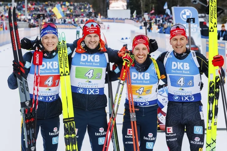 Vier Biathleten in Wettkampfausrüstung halten Skier, lächelnd vor einer Winterkulisse.