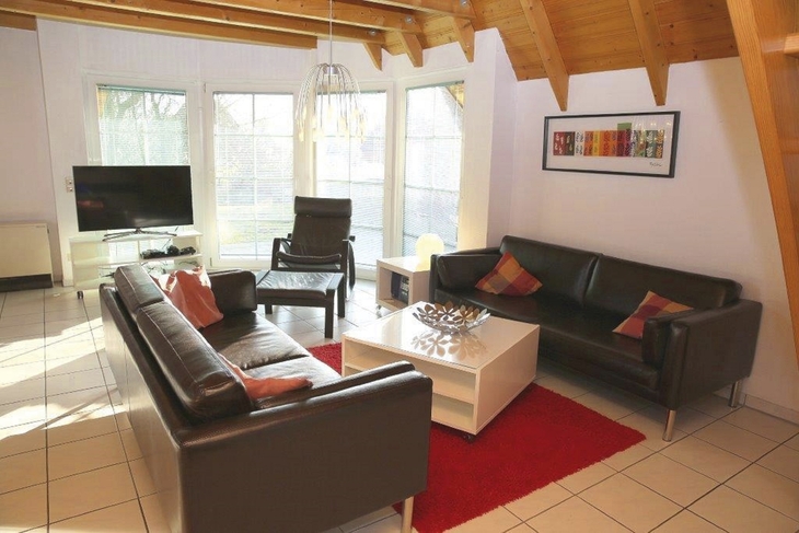 Wohnzimmer mit zwei Sofas, Sessel, Couchtisch, Fernseher und großen Fenstern.