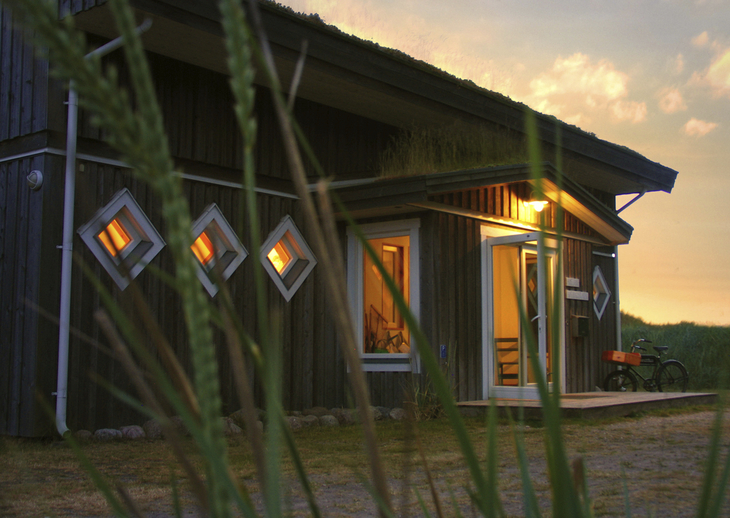 Holzhaus mit leuchtenden Fenstern bei Sonnenuntergang, ein Fahrrad steht davor.