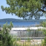 Blick auf einen See durch Kiefern und Sanddünen mit Holzzaun im Vordergrund.