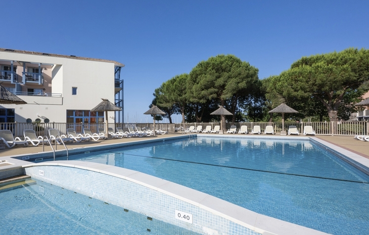 Poolbereich einer Ferienresidenz mit Sonnenliegen und Bäumen im Hintergrund.