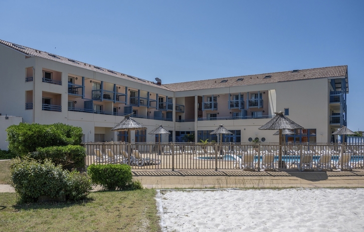 Gebäude mit Pool, Sonnenschirmen und Liegestühlen, blauer Himmel im Hintergrund.