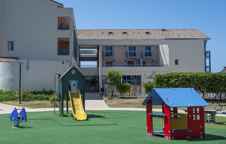 Spielplatz mit Rutsche und Spielhäusern vor einem Gebäude.