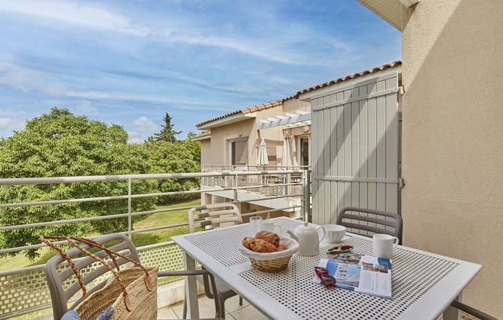 Balkon mit Tisch, Frühstück, Korb, Aussicht auf Grünfläche und Wohnhäuser im Hintergrund.
