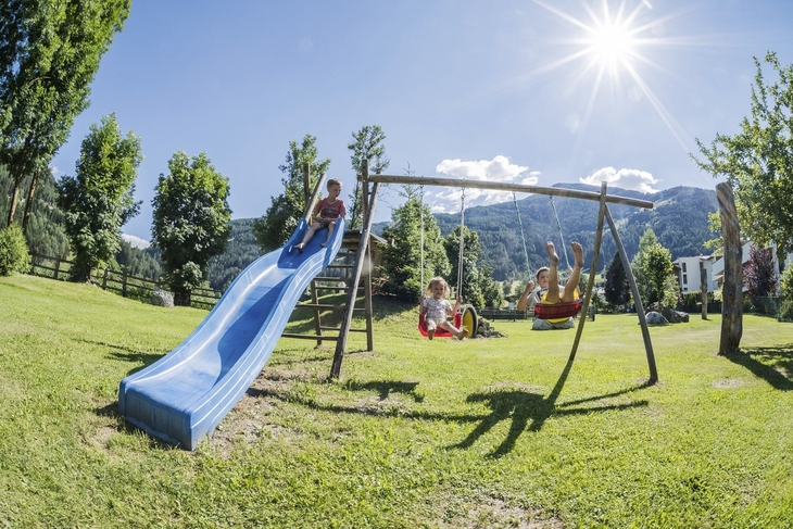Kinder spielen auf einem Spielplatz mit Rutsche und Schaukel bei Sonnenschein.