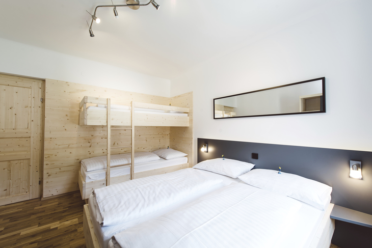 Zimmer mit Doppelbett und Etagenbett, helle Holzwände, moderne Beleuchtung.