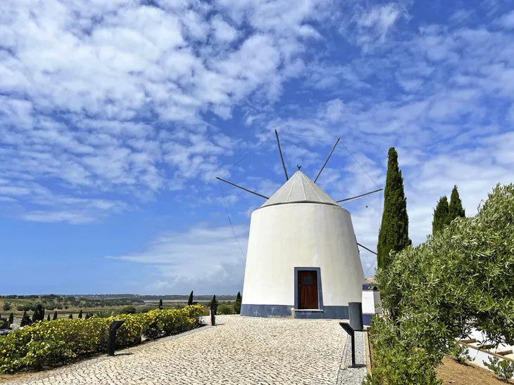 Windmühle in Vila Real, Algarve, umgeben von Flora und blauem Himmel.