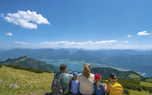 Familie sitzt auf Wiese mit Blick auf See und Berge unter blauem Himmel.