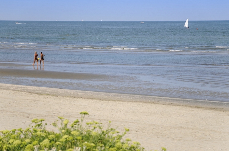 Strand mit Spaziergängern am Meer, im Hintergrund ein Segelboot.