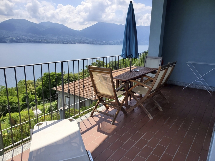 Terrasse mit Tisch, Stühlen und Schirm, Blick auf See und Berge im Hintergrund.