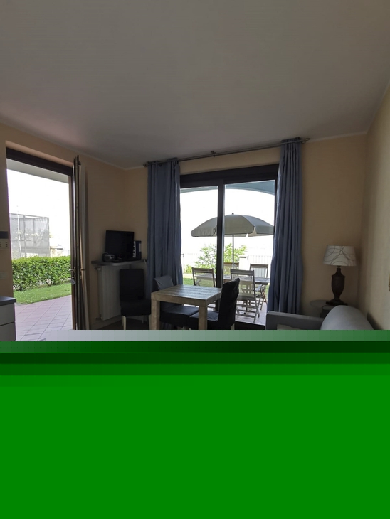 Wohnzimmer mit grauem Sofa, Tisch, großer Tür zum Balkon mit Blick auf Garten und Sonnenschirm.