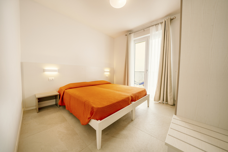 Helles Schlafzimmer mit Doppelbett, oranger Bettdecke und Zugang zu einem Balkon.