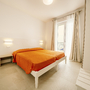 Helles Schlafzimmer mit Doppelbett, oranger Bettdecke und Zugang zu einem Balkon.