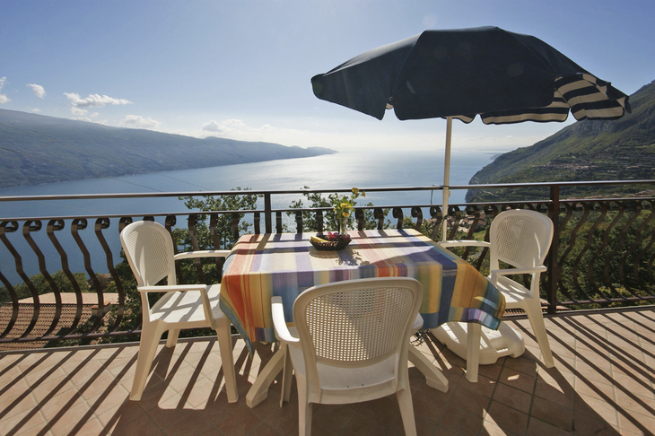 Balkon mit Tisch und Stühlen, Blick auf See und Berge, blauer Sonnenschirm.
