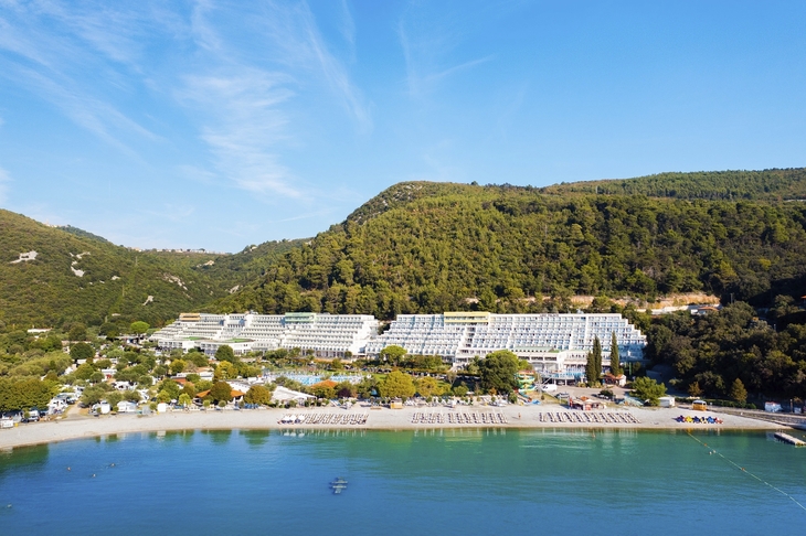 Hotel am Strand mit grünen Hügeln im Hintergrund und blauem Himmel.