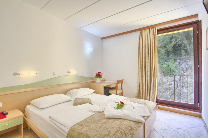 Doppelzimmer mit Doppelbett, Tisch, Fenster mit Vorhang, Blick auf die Straße, helle Einrichtung.