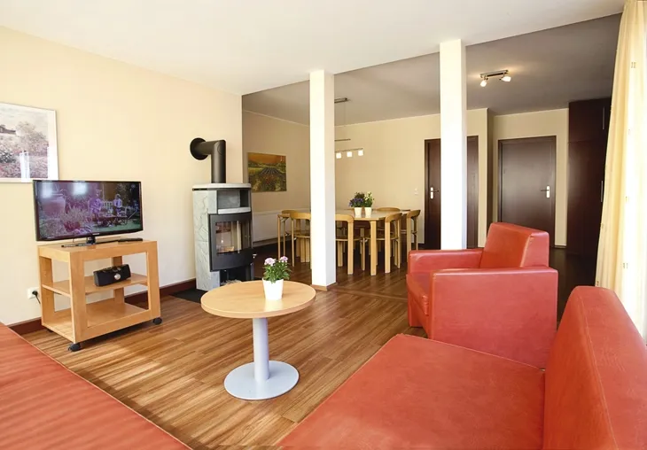 Wohnzimmer mit rotem Sofa, kleinem Tisch, Ofen, Fernseher und Esstisch mit Stühlen in der Ferienanlage Prüm.