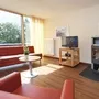 Wohnzimmer in der Ferienanlage Prüm, mit rotem Sofa und Sessel, kleiner Tisch, Holzofen, Fernseher und Blick auf Bäume durch große Fenster.