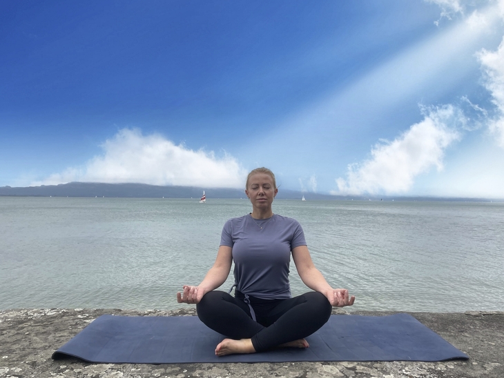 Person meditiert im Lotussitz auf einer Yogamatte am Ufer eines Sees.