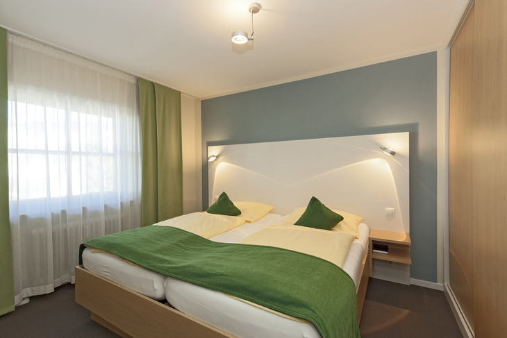 Hotelzimmer mit Doppelbett, grüner Bettdecke und hellem Holzmobiliar.