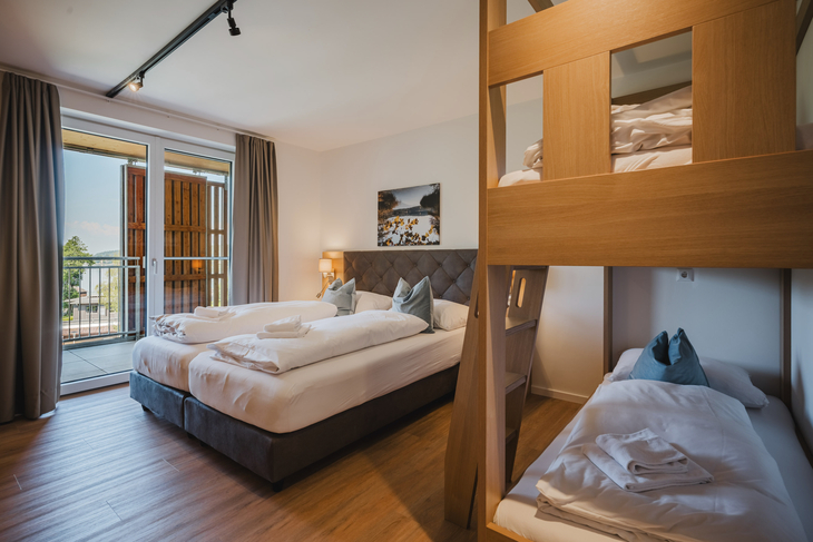 Hotelzimmer mit Doppelbett, Etagenbett, Balkon und moderner Einrichtung.
