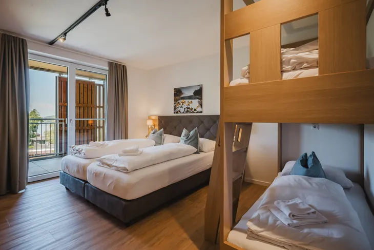 Hotelzimmer mit Doppelbett, Etagenbett, Balkon und moderner Einrichtung.