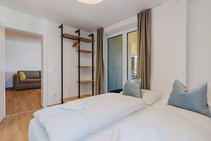 Helles Schlafzimmer mit Doppelbett, offenem Regal und Zugang zum Balkon.