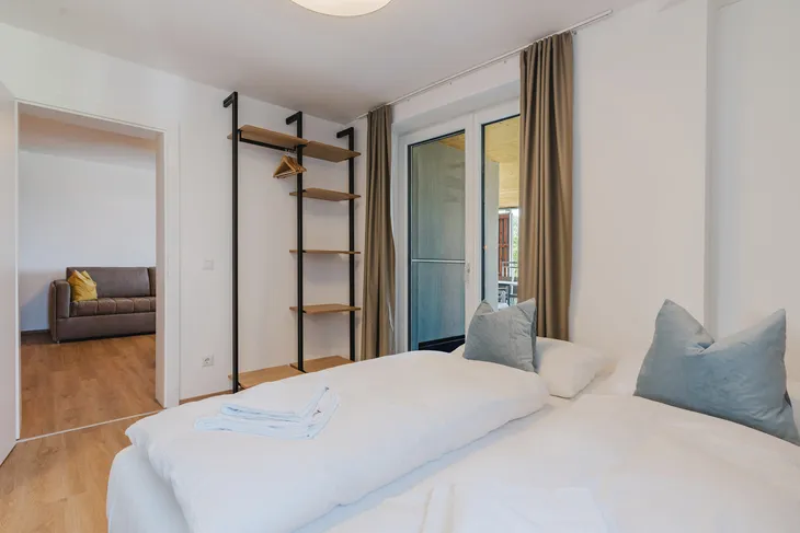 Helles Schlafzimmer mit Doppelbett, offenem Regal und Zugang zum Balkon.