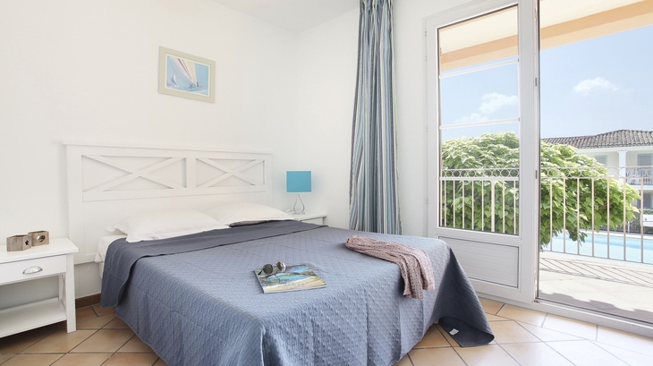 Helles Schlafzimmer mit Doppelbett, Meeresbild, Lampe, Balkonblick auf Pool und Bäume.