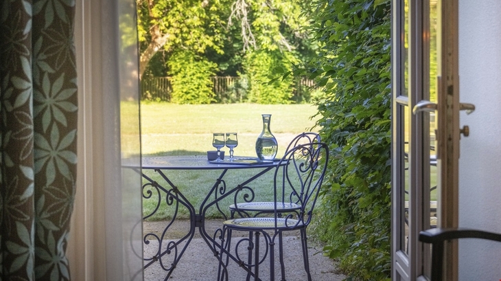 Metalltisch mit zwei Stühlen im Garten mit Blick durch offene Tür.