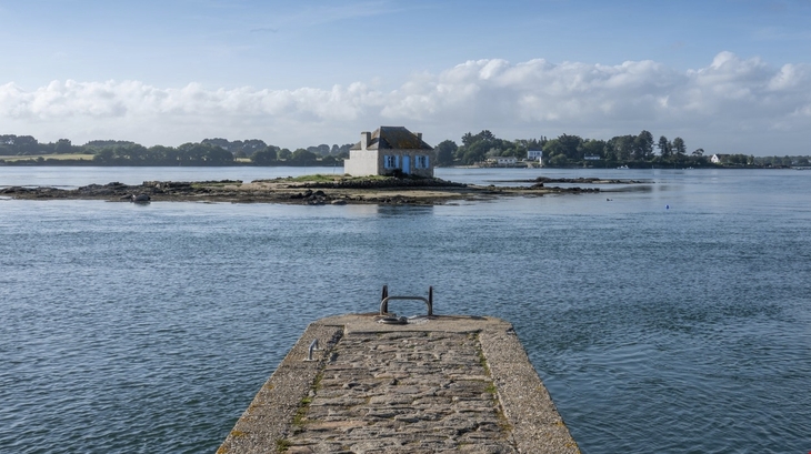 Steg führt zu einer kleinen Insel mit Haus im ruhigen Wasser unter blauem Himmel.