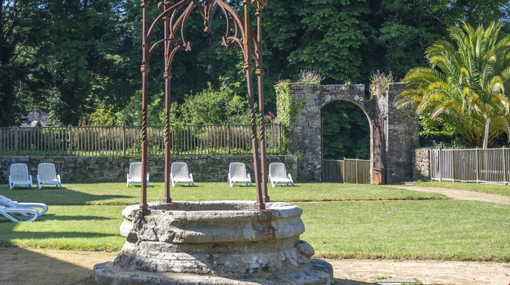 Steinbrunnen im Garten vor einer Mauer mit Torbogen und Liegestühlen.