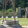 Steinbrunnen im Garten vor einer Mauer mit Torbogen und Liegestühlen.