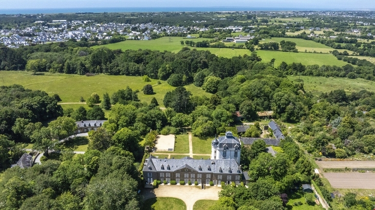 Luftaufnahme eines Schlosses in grüner Landschaft mit Meer im Hintergrund.