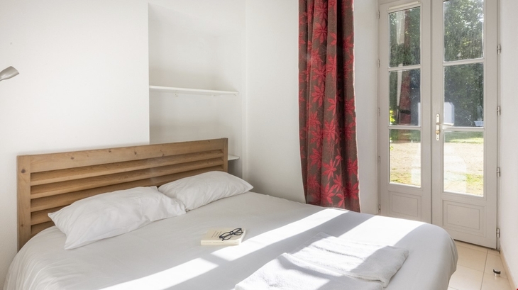 Helles Schlafzimmer mit Doppelbett, roten Vorhängen und Sonnenlicht durch Terrassentür.
