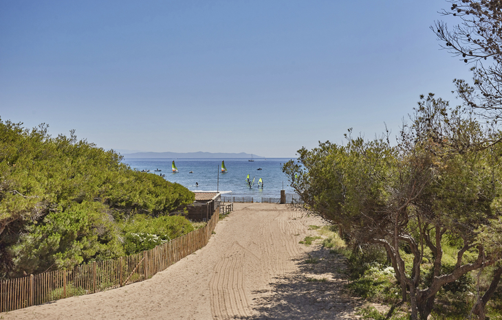 Sandweg führt zum Strand mit Bäumen und segelnden Windsurfern im Hintergrund.