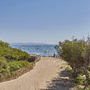 Sandweg führt zum Strand mit Bäumen und segelnden Windsurfern im Hintergrund.