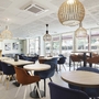 Moderne Café-Lounge mit Holztischen, blauen und braunen Stühlen und großen Fenstern.