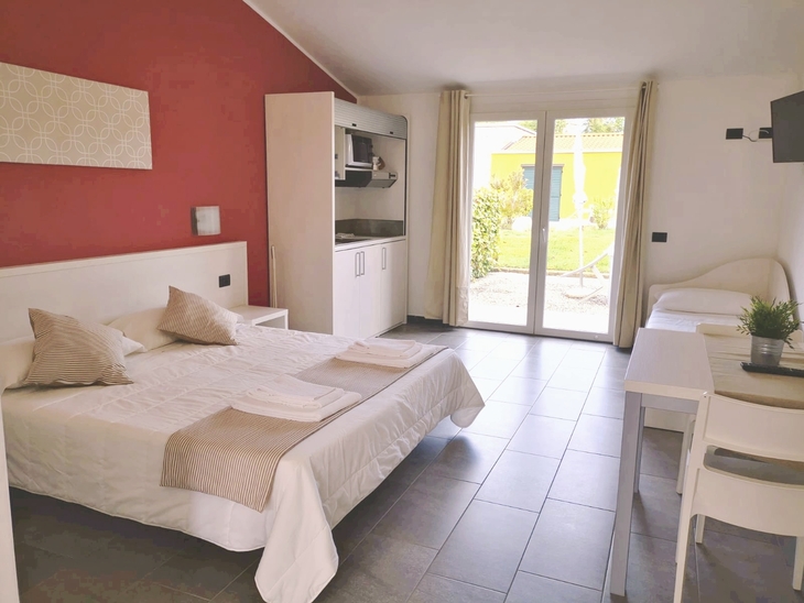 Hotelzimmer mit Bett, Tisch, Küchenzeile und Fenster mit Blick auf Garten.