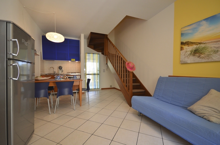Wohnbereich mit blauer Couch, Esstisch, Küche und Holztreppe in modernem Apartment.