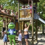 Kinder und Maskottchen auf einem Spielplatz mit Klettergerüst und Rutsche.