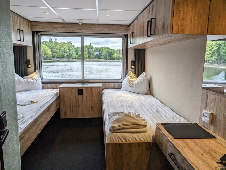 Zwei-Bett-Kabine auf dem Oberdeck eines Schiffes mit Fensterblick aufs Wasser.