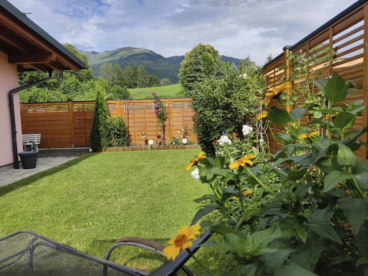 Garten eines Bungalows mit Blumen, Holzzaun und Bergblick im Hintergrund.