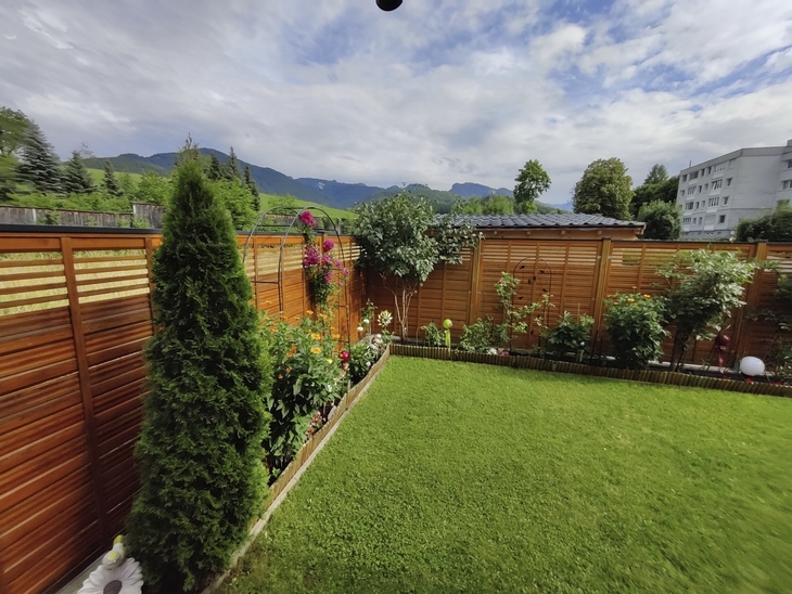Garten mit grünem Rasen, Holzsichtschutz, Blumen, Bergen im Hintergrund, wolkiger Himmel.
