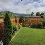 Garten mit grünem Rasen, Holzsichtschutz, Blumen, Bergen im Hintergrund, wolkiger Himmel.