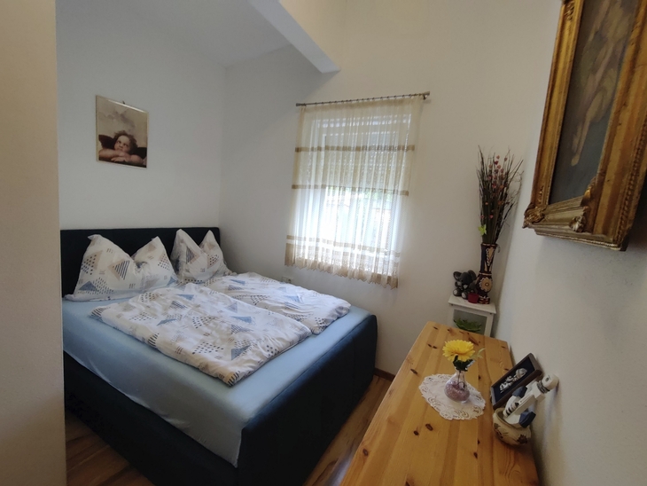 Kleines Schlafzimmer mit Doppelbett, Holztisch, Bild an der Wand und Tageslichtfenster.